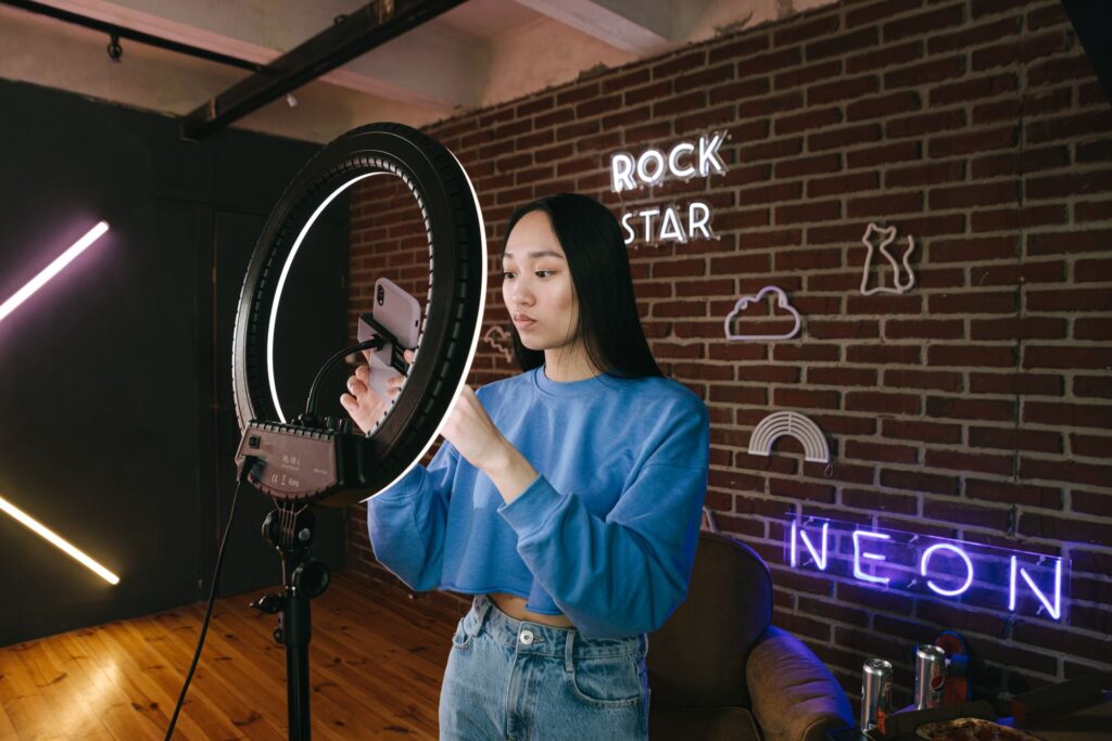 Woman adjusting her phone on a ring light stand in a studio with brick walls and neon signs.